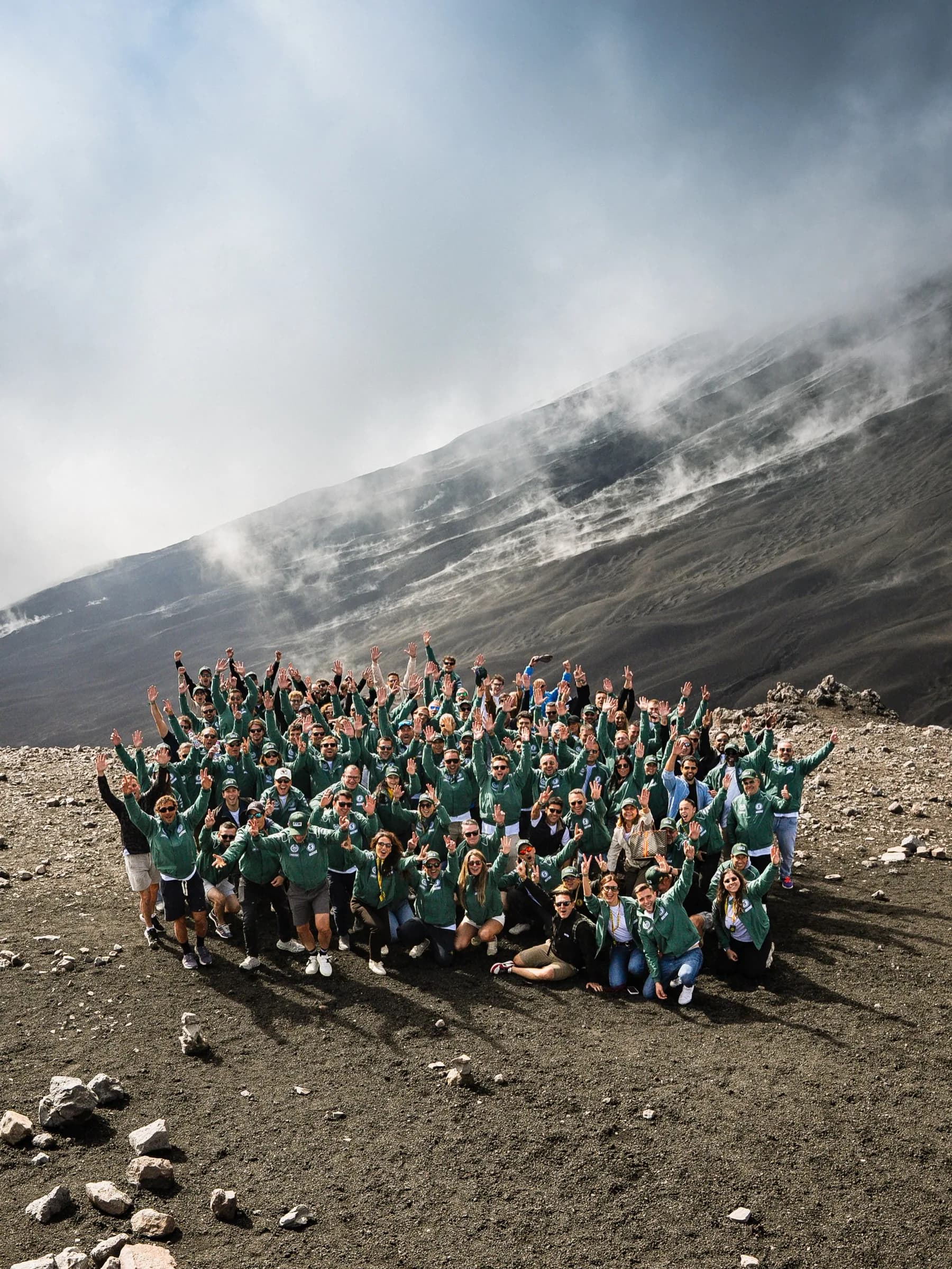 Around a hundred people in SIX2SIX Driver jackets, arms raised, on the volcanic slope of Etna under cloud