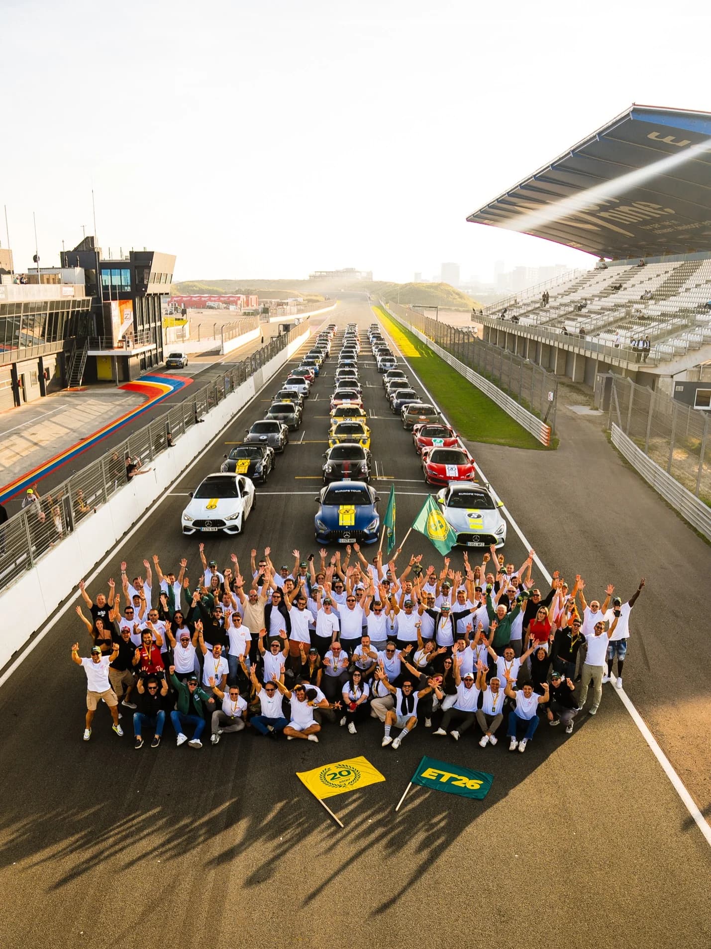 SIX2SIX drivers in white tour t-shirts on the starting grid at Zandvoort, three rows of supercars behind them, the yellow 20 and green ET26 flags on the tarmac
